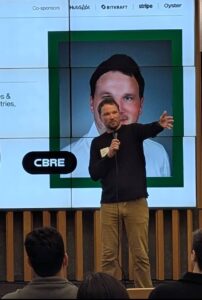 A speaker shown near a large screen at an event.